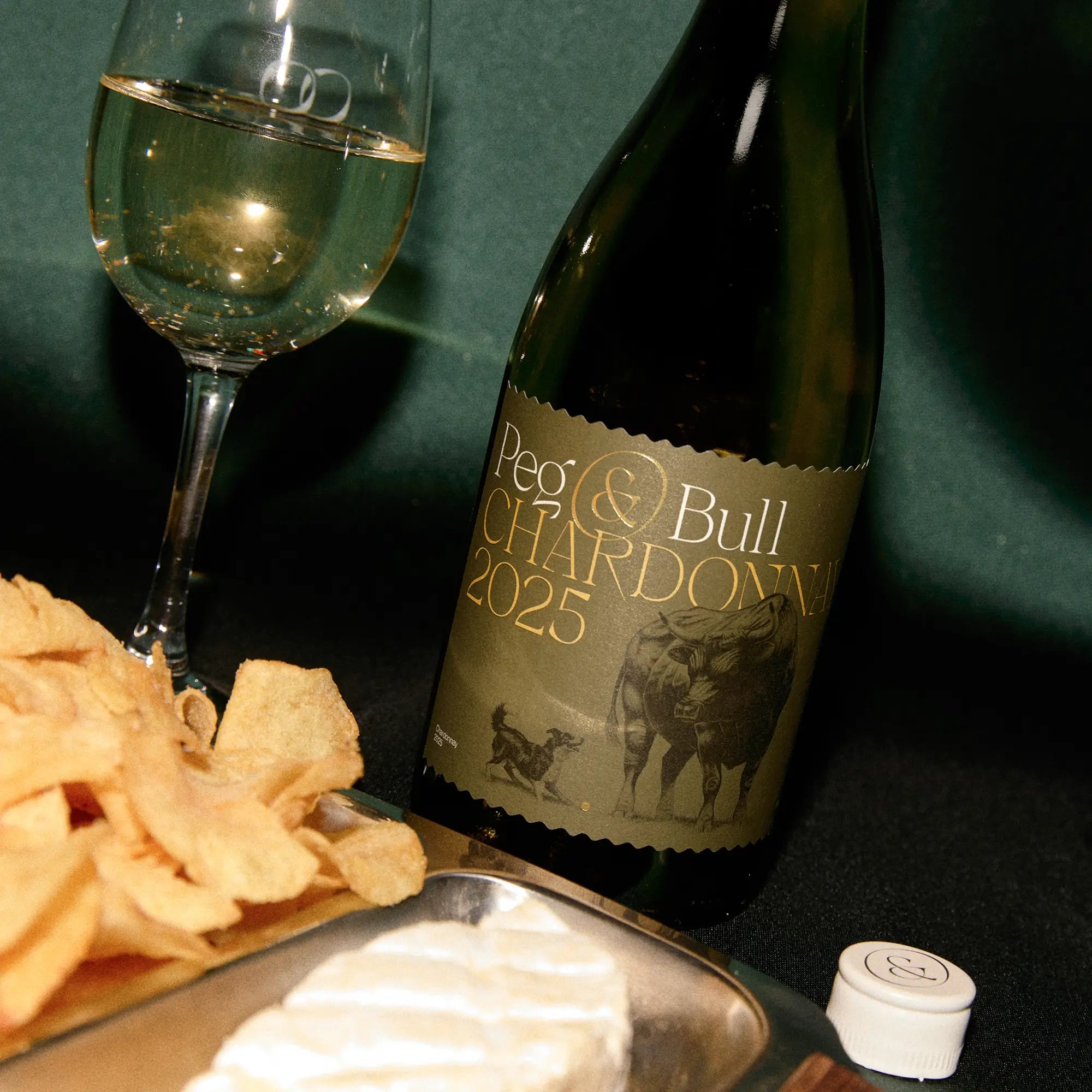 Bottle of Peg & Bull Chardonnay 2025 with a glass of wine and snacks on a dark surface
