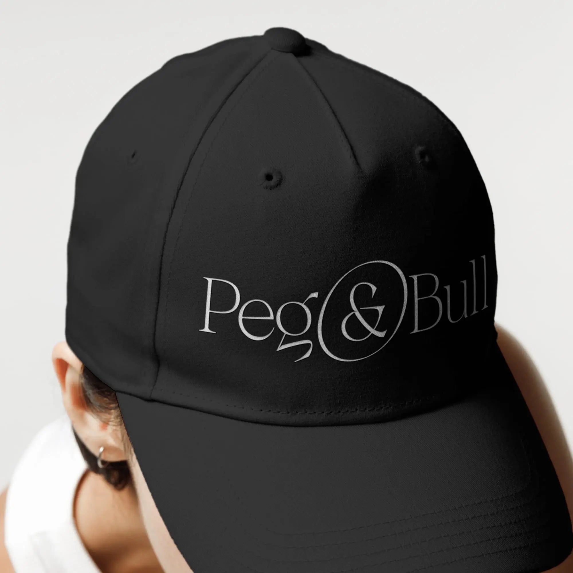 Black cap with 'Peg & Bull' logo worn by a person on a light gray background
