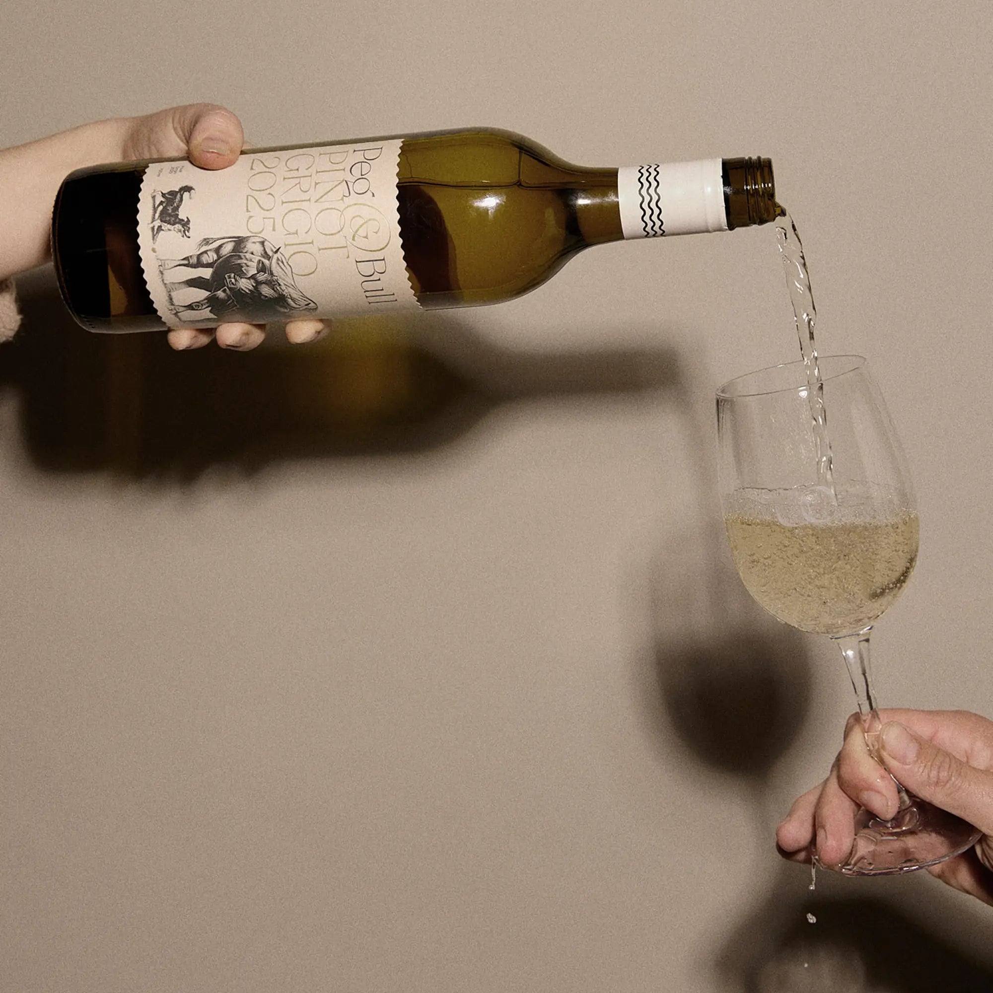 Person pouring white wine from a bottle of Peg & Bull Pinot Grigio, 2025 into a glass against a neutral background