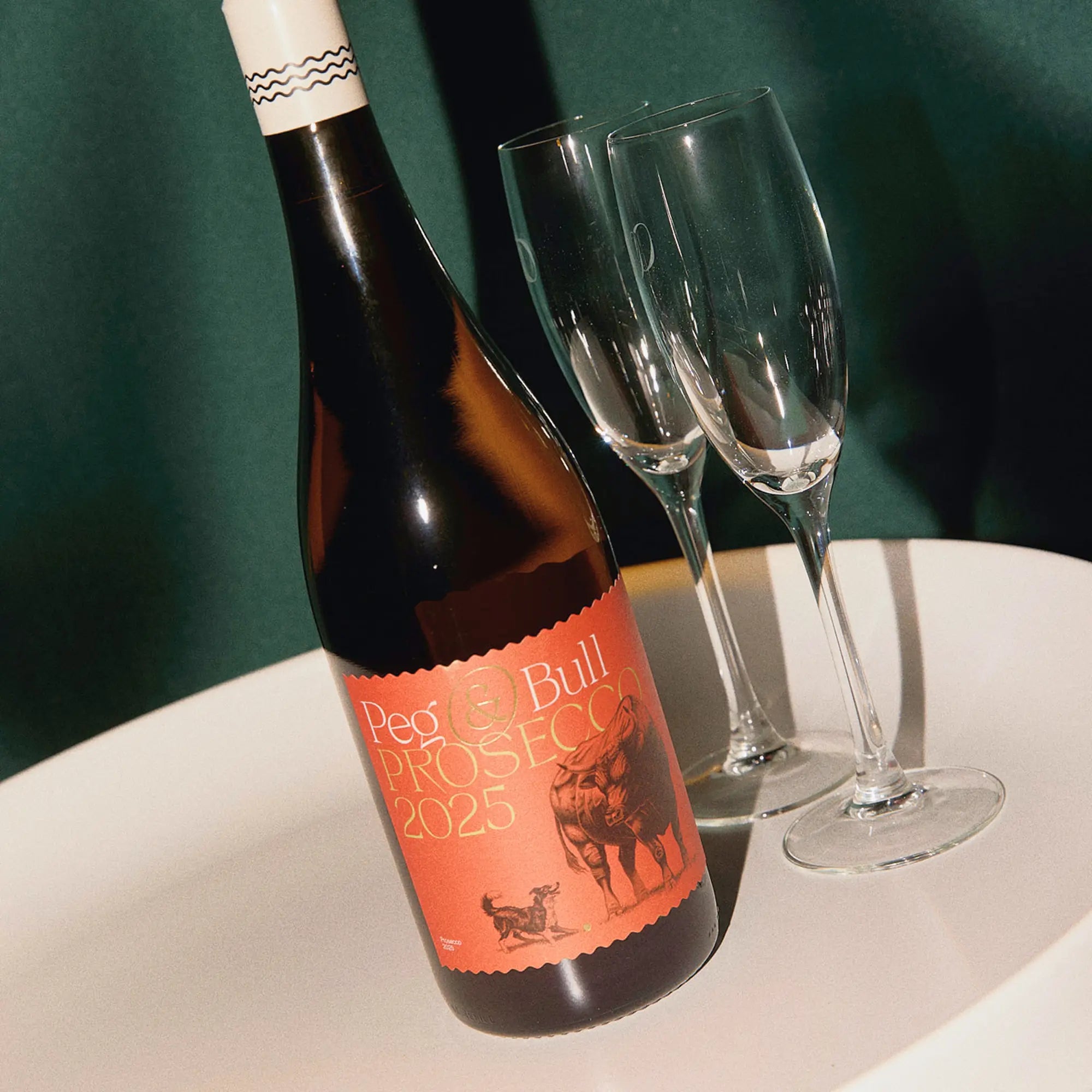 Bottle of 'Peg's Bull Prosecco 2025' with two empty glasses on a white tray against a dark green background.