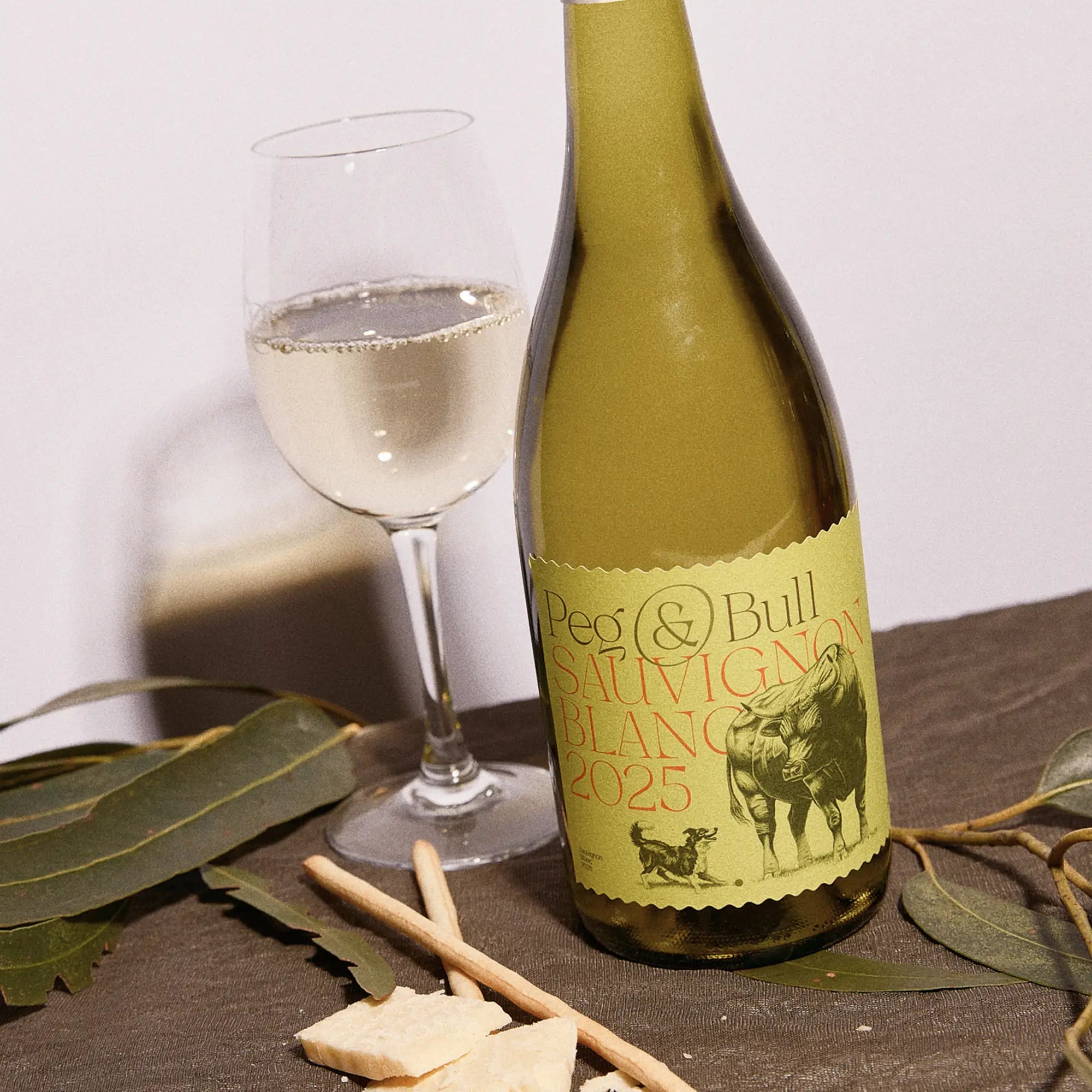 Peg & Bull Sauvignon Blanc 2025 wine bottle with a glass of white wine and some snacks on a table.