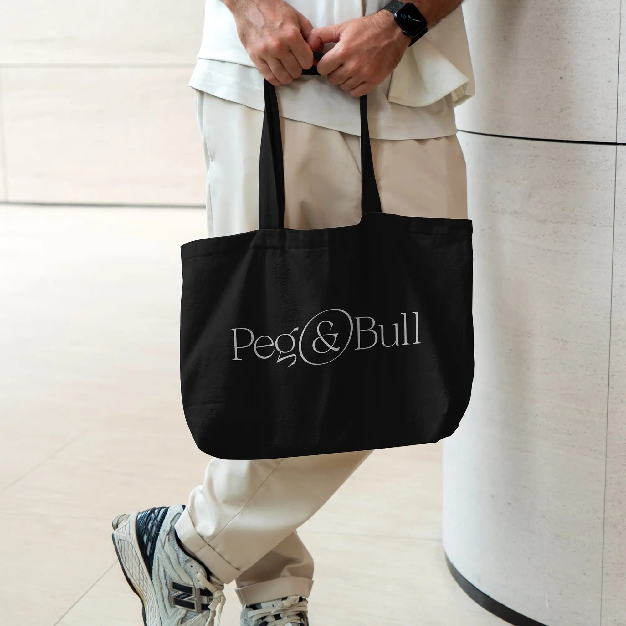Person holding a black tote bag with 'Peg & Bull' logo against a light background